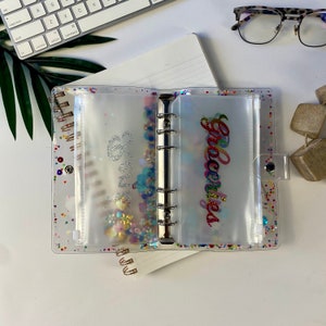 May include: Clear, glitter-filled binder with pockets labeled "Savings" and "Groceries" in cursive. The binder has a silver metal ring mechanism and a clear snap closure. The pockets are filled with colorful confetti and decorative elements.
