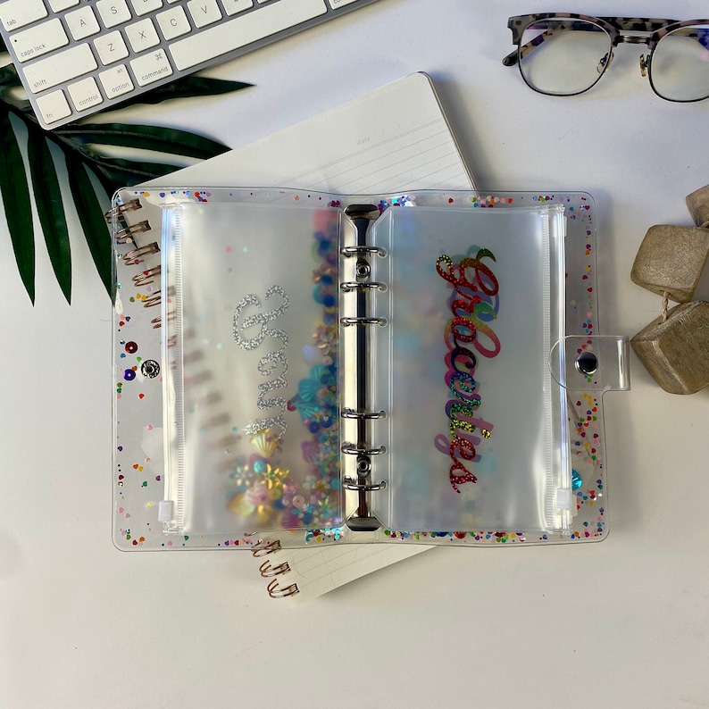 May include: Clear, glitter-filled binder with pockets labeled "Savings" and "Groceries" in cursive. The binder has a silver metal ring mechanism and a clear snap closure. The pockets are filled with colorful confetti and decorative elements.