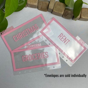 May include: Clear plastic budget envelopes with pink borders and labels. The labels read "CHRISTMAS", "GROCERIES", and "RENT" in pink lettering. The text "*Envelopes are sold individually" is at the bottom.