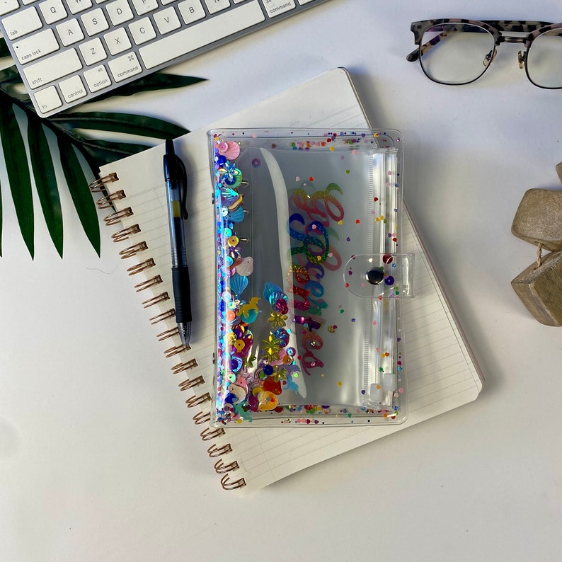 May include: A clear, plastic planner with colorful sequins and the word "Spectacular" in script. The planner is closed with a clear snap and rests on a spiral notebook. A black pen and a keyboard are also visible.