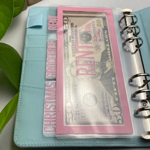 May include: A light blue binder with silver rings, holding clear plastic envelopes. One envelope contains a fifty-dollar bill and the word "RENT" in pink. Another envelope has the words "CHRISTMAS GROCERIES" in pink.