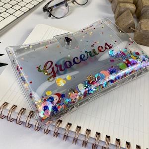 May include: A clear plastic Groceries wallet with a snap closure. The wallet is filled with colorful sequins and confetti. The word "Groceries" is written in rainbow script across the front. The wallet is on top of a spiral notebook.