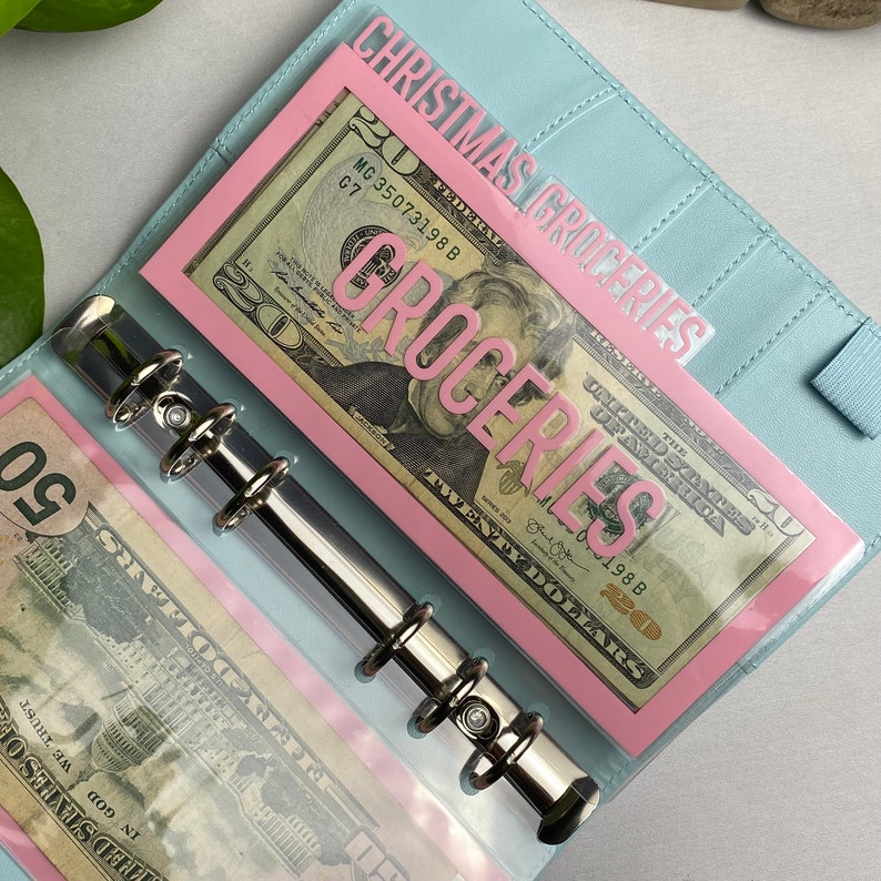 May include: A light blue binder with a silver metal ring mechanism. Inside, a pink divider reads "CHRISTMAS GROCERIES" and "GROCERIES" over a twenty-dollar bill. Another pocket contains a fifty-dollar bill. Budgeting and organization.