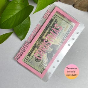 May include: A pink and clear budget envelope with the words "CHRISTMAS GROCERIES" and "CHRISTMAS" printed on it. A twenty-dollar bill is visible inside the envelope. The word "RENT" is also printed on the envelope.