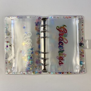 May include: Clear, glitter-filled binder with pockets labeled "Bills" and "Groceries" in cursive. The binder has a silver ring mechanism and a snap closure. The pockets are filled with colorful confetti.