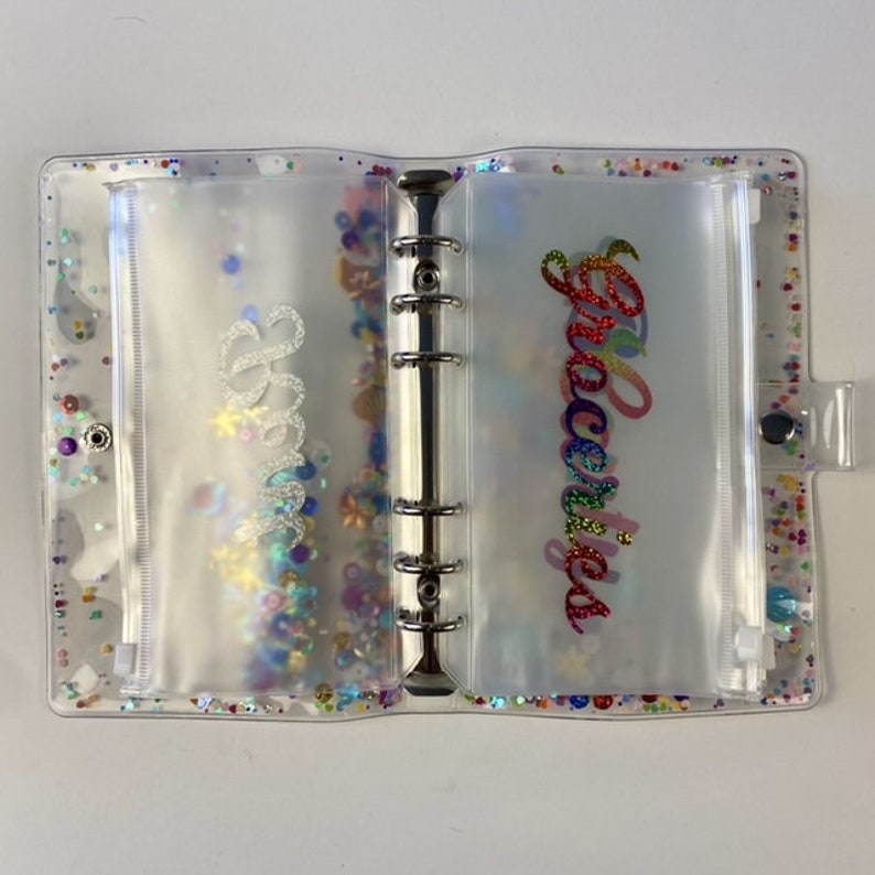 May include: Clear, glitter-filled binder with pockets labeled "Bills" and "Groceries" in cursive. The binder has a silver ring mechanism and a snap closure. The pockets are filled with colorful confetti.