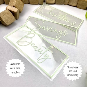 May include: Clear plastic budget envelopes with a light green border and script lettering. The envelopes are labeled "Christmas", "Savings", and "Beauty". The text "Available with Hole Punches" and "*Envelopes are sold individually" are also visible.