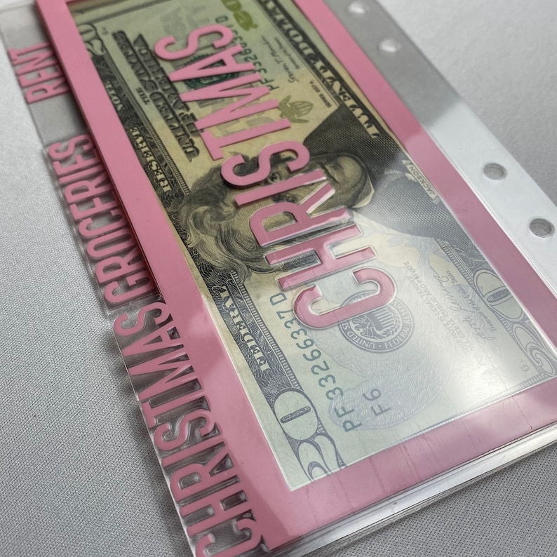 May include: A clear plastic budget envelope with pink accents and the word "CHRISTMAS" in pink lettering. The envelope contains a twenty-dollar bill. The words "GROCERIES" and "RENT" are also visible.
