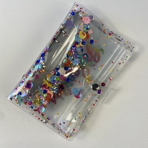 May include: Clear, rectangular binder with colorful sequins and embellishments. The binder features a snap closure and silver metal rings. The interior is visible, and the design includes floral and seashell shapes, with the word "Aloha" visible.