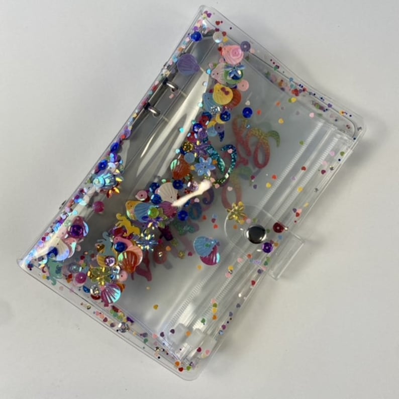 May include: Clear, rectangular binder with colorful sequins and embellishments. The binder features a snap closure and silver metal rings. The interior is visible, and the design includes floral and seashell shapes, with the word "Aloha" visible.