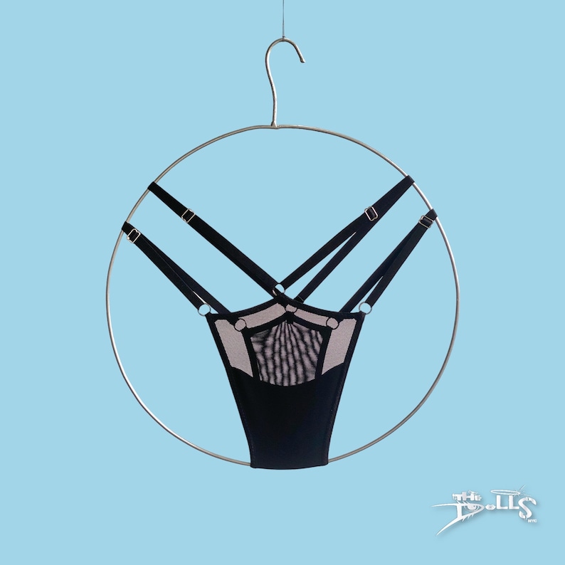 May include: Black thong underwear featuring a sheer mesh panel at the front, multiple adjustable elastic straps with silver-toned metal rings and sliders, all suspended from a circular metal hanger against a light blue background.