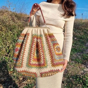 Könnte beinhalten: Eine große, cremefarbene Häkeltasche mit einem farbenfrohen geometrischen Muster. Die Tasche hat einen Metallverschluss und braune Ledergriffe. Das Häkeldesign enthält Quadrate in verschiedenen Farben, darunter Rosa, Gelb, Grün und Blau.