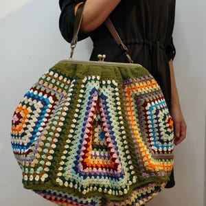 May include: A large, handmade crochet bag with a colorful granny square design. The bag features a green border and a metal clasp closure. Brown leather handles and a shoulder strap are attached to the bag.