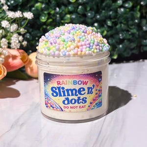 May include: A transparent plastic jar filled with white slime and topped with pastel-coloured foam beads. The jar has a label that reads "RAINBOW Slime n' dots" with the words "DO NOT EAT" below.
