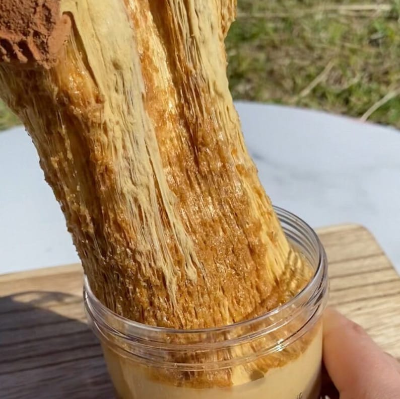 May include: A close-up of a brown and beige slime-like substance being pulled out of a clear glass jar. The slime is very stretchy and has a grainy texture.