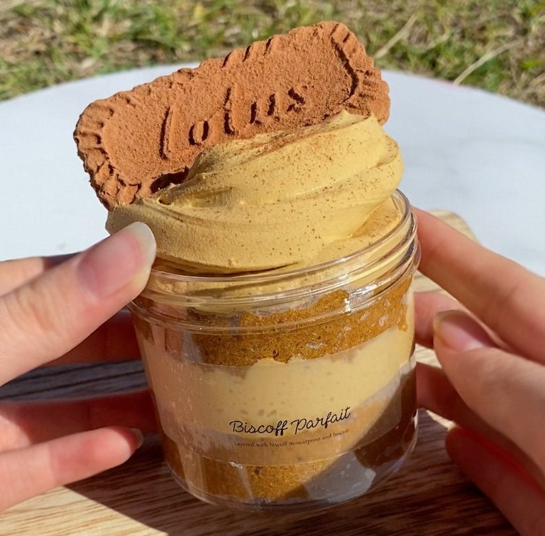 May include: A glass jar filled with layers of brown and beige dessert topped with a brown cookie with the word "Lotus" printed on it. The label on the jar reads "Biscoff Parfait Layered with Biscoff mascarpone and biscuit".