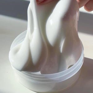 May include: A thick, glossy, opaque white slime substance being pulled upwards from a clear, round plastic container. The material has a smooth, viscous texture that stretches and drips back into the jar, reflecting light on its surface.