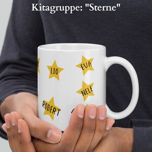 May include: White ceramic mug featuring yellow star graphics with names in black text. The mug has a handle and the text "Kitagruppe: Sterne" is at the top. The mug is held by a person.