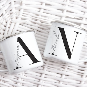 Op de afbeelding: Twee witte emaille mokken met zwarte handvatten. De mokken hebben een zwarte letter gedrukt op de voorkant, "A" op de ene mok en "N" op de andere. De mokken staan op een wit rieten oppervlak. De tekst op de mokken luidt "Antonia" en "Nicole".
