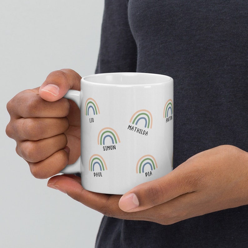 May include: White ceramic mug featuring colorful rainbow designs with names like "Lio," "Simon," and "Mathilda." The rainbows are in pastel shades of blue, green, and peach. The mug is held by a person.