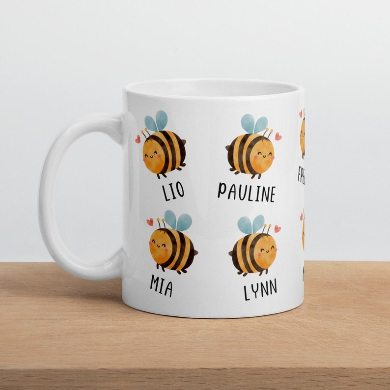 May include: White ceramic mug featuring cartoon bees with names like Lio, Pauline, Mia, and Lynn. Each bee has a smiling face, wings, and a heart. The mug is on a wooden surface.