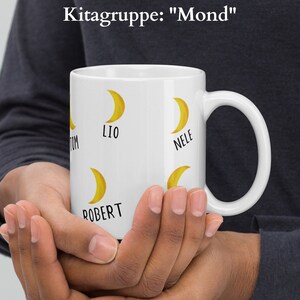 May include: White ceramic mug featuring yellow crescent moon graphics and names. The mug has the text "Kitagruppe: Mond" at the top. The names Tom, Lio, Nele, and Robert are printed on the mug. The mug is held in both hands.