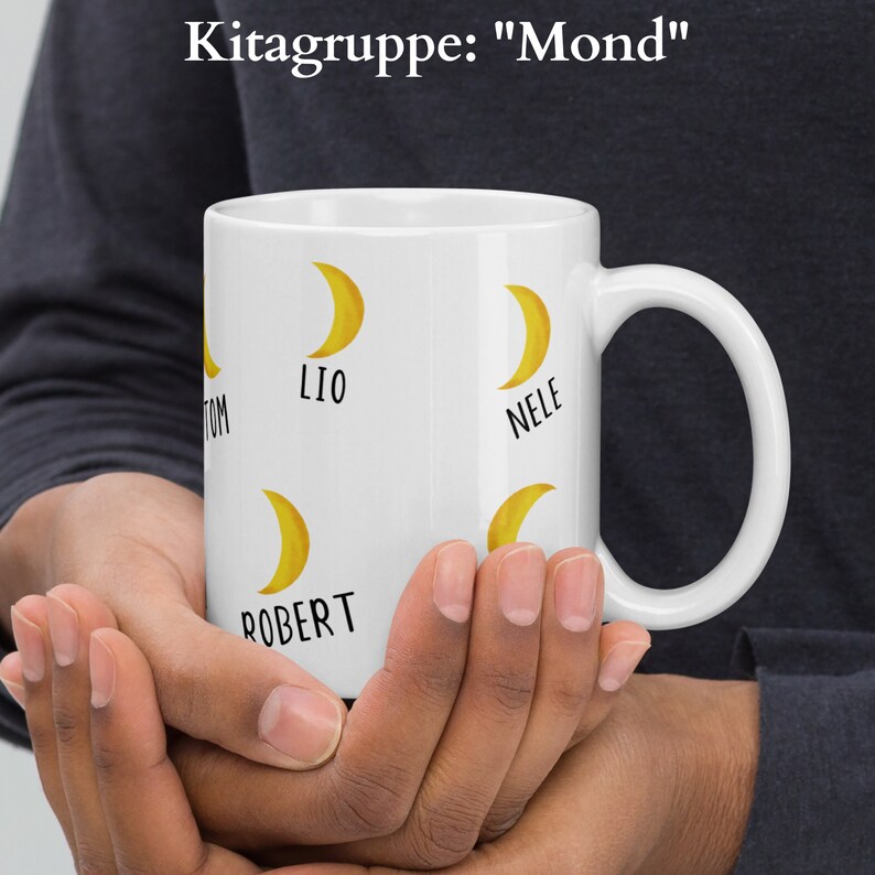 May include: White ceramic mug featuring yellow crescent moon graphics and names. The mug has the text "Kitagruppe: Mond" at the top. The names Tom, Lio, Nele, and Robert are printed on the mug. The mug is held in both hands.