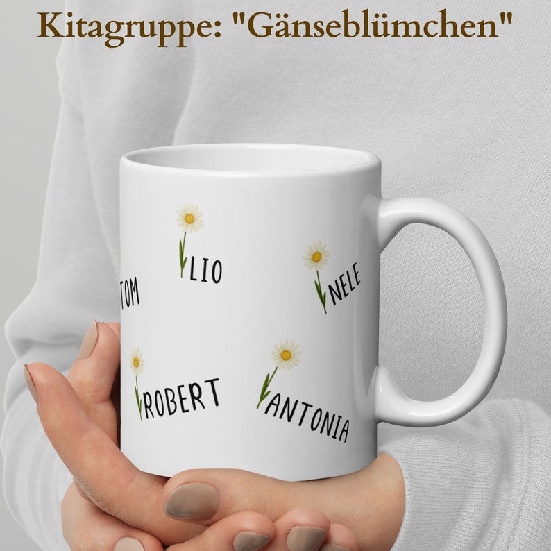May include: White ceramic mug featuring daisy illustrations and names: TOM, LIO, NELE, ROBERT, and ANTONIA. The mug has a curved handle and the text "Kitagruppe: "G&auml;nsebl&uuml;mchen"" at the top. The mug is held by a person.