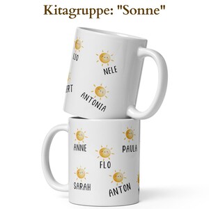 May include: Two white ceramic mugs stacked on top of each other. Each mug features a repeating pattern of smiling sun illustrations and names. The text "Kitagruppe: Sonne" is at the top. The mugs have a handle on the side.