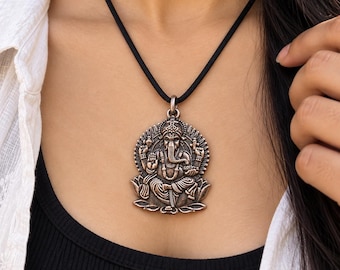 Puede incluir: Un colgante metálico con un relieve detallado de la deidad Ganesha sentada sobre una flor de loto, suspendido de un cordón negro sencillo. El colgante tiene un acabado oxidado que resalta las intrincadas tallas de la figura y el halo circundante.