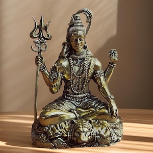 Pode incluir: Uma est&aacute;tua de metal em tom dourado da divindade Shiva sentada em posi&ccedil;&atilde;o meditativa sobre uma pele de tigre. A figura segura um tridente com um s&iacute;mbolo Om, possui m&uacute;ltiplos bra&ccedil;os, usa colares de contas e uma lua crescente no cabelo.