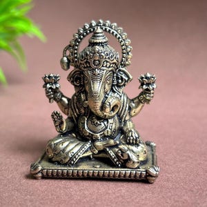 May include: A gold-colored metal statue of Ganesha, the Hindu god of beginnings, sitting on a square base. The statue is intricately detailed with a crown, a trunk, and four arms.