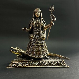 K&ouml;nnte beinhalten: Detaillierte Bronzestatue einer weiblichen Figur, geschm&uuml;ckt mit aufwendigem Schmuck und einem gemusterten Kleid, das einen Dreizack h&auml;lt. Die Figur steht auf einem Krokodil auf einem dekorativen Sockel. Die Statue hat eine dunkelbronze Oberfl&auml;che.