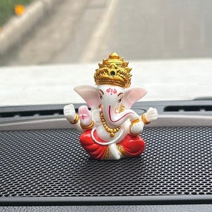 Könnte beinhalten: Eine kleine weiße Figur des hinduistischen Gottes Ganesha, der mit gekreuzten Beinen sitzt und ein kleines Objekt in seiner rechten Hand hält. Er trägt eine rote und goldene Robe und eine goldene Krone mit einem grünen Juwel. Die Figur sitzt auf einem schwarzen Lautsprechergitter in einem Auto.