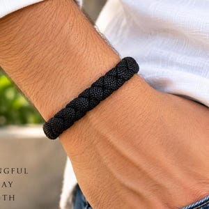 May include: A black braided cord bracelet featuring a thick, woven texture. The text on the left side of the image reads: SIMPLE MEANINGFUL EVERYDAY STRENGTH. The bracelet is worn on a wrist, showcasing the dark, uniform color and intricate knotting.