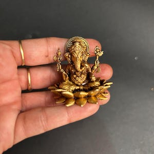 May include: A small, metallic gold-toned figurine of the deity Ganesha, depicted with an elephant head and four arms, seated in a cross-legged position upon a stylized lotus flower base with detailed petal carvings and an ornate headdress.