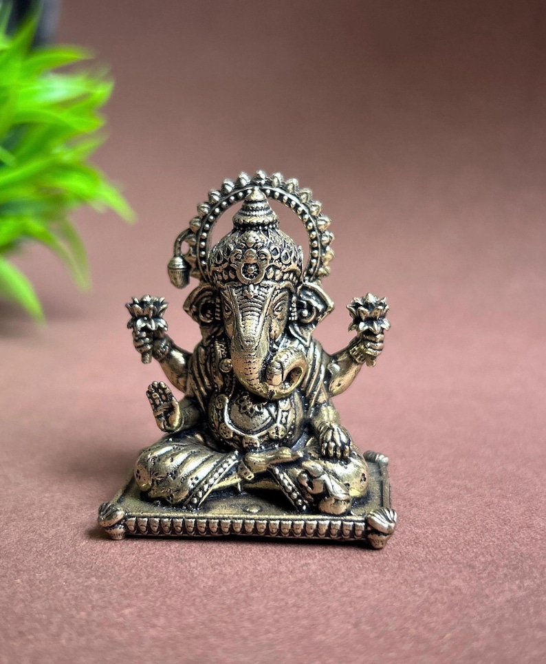 May include: A gold-colored metal statue of Ganesha, the Hindu god of beginnings, sitting on a square base. The statue is intricately detailed with a crown, a trunk, and four arms.