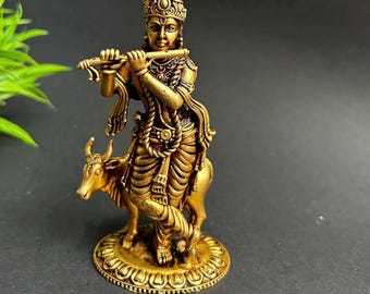 May include: A gold-coloured statue of a deity playing a flute, standing on a cow. The statue has intricate details, including ornate clothing and a detailed crown. The base of the statue is round with a decorative border. The background is dark.