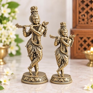 May include: Two brass-coloured statues of a deity playing a flute, each standing on a circular base with intricate patterns. The figures are adorned with detailed jewellery, crowns, and flowing garments, with a small bird perched on the flute.