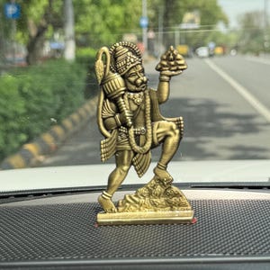 May include: A bronze-colored statue of a Hindu deity, Hanuman, holding a mace and a mountain. The statue is detailed with intricate carvings and stands on a small base. The background shows a road and trees.