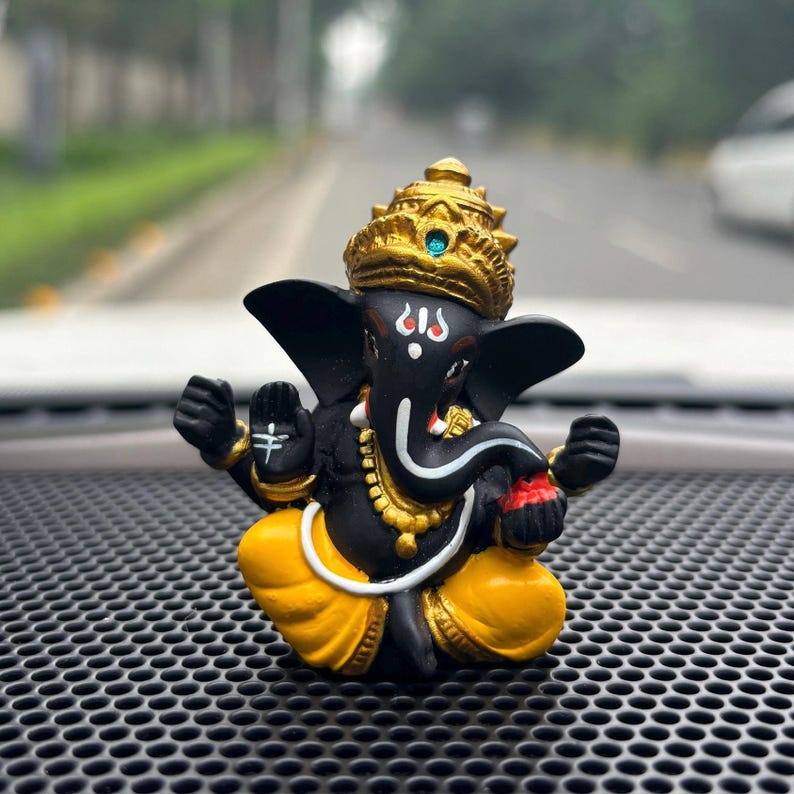 Puede incluir: Una figurita negra y dorada del dios hind&uacute; Ganesha. La figurita est&aacute; sentada con las piernas cruzadas y lleva un dhoti amarillo. La figurita lleva una corona dorada con una joya azul en la parte superior. La figurita est&aacute; sentada sobre una superficie negra con un patr&oacute;n de panal.