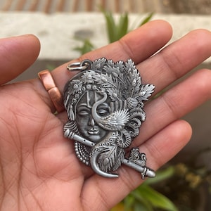 May include: Silver pendant featuring a detailed depiction of Krishna playing a flute, with a peacock feather design and a peacock behind him.