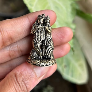 Puede incluir: Una figurita de metal dorado de Radha y Krishna de pie juntos. Krishna sostiene una flauta y Radha tiene su brazo alrededor de él. La figurita está sobre una base con un diseño floral.