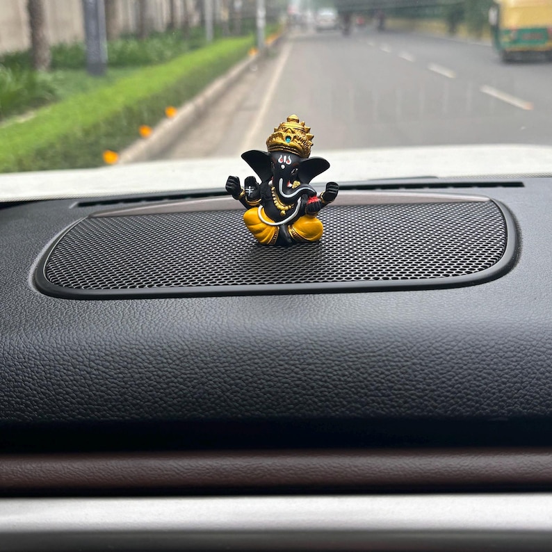 Puede incluir: Una figurita de Ganesha negra y dorada sentada en el tablero de un coche negro. La figurita est&aacute; en posici&oacute;n sentada con las manos en mudra.