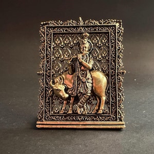 May include: A metallic gold-toned rectangular plaque featuring a relief sculpture of a figure playing a flute while standing beside a cow. The central design is enclosed within an ornate, filigree-style frame with intricate floral patterns.