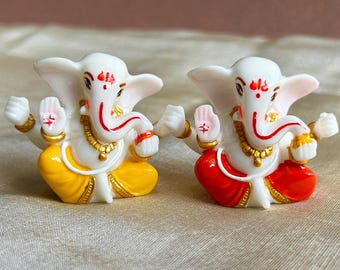 May include: Two small white Ganesha figurines, one with a yellow dhoti and the other with a red dhoti. Both figurines have gold accents and red and white painted details.