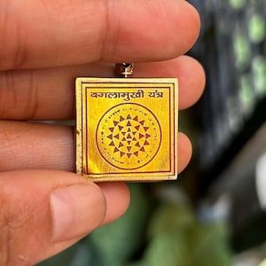 May include: A gold-colored pendant with a red and gold design and the text "कालामुर्ती यंत्र" in Sanskrit.