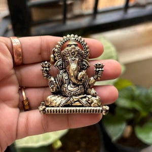 May include: A gold-toned metal pendant featuring a detailed depiction of the Hindu deity Ganesha. The pendant shows Ganesha sitting on a throne with his trunk raised and holding a lotus flower in each hand.