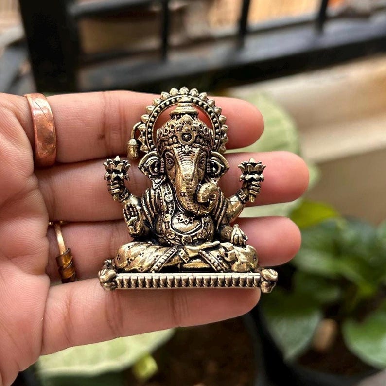 May include: A gold-toned metal pendant featuring a detailed depiction of the Hindu deity Ganesha. The pendant shows Ganesha sitting on a throne with his trunk raised and holding a lotus flower in each hand.