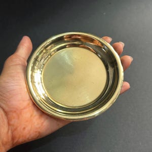 Könnte beinhalten: Eine kleine, runde, messingfarbene Schale mit leicht erhöhtem Rand. Die Schale hat eine glatte, reflektierende Oberfläche und wird in einer Hand vor dunklem Hintergrund gehalten. Die Schale scheint aus Metall zu sein.
